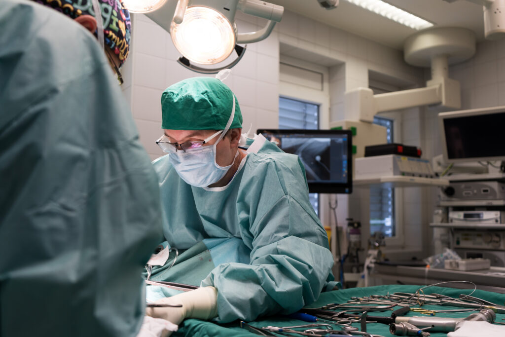 Zwei Personen stehen in einem OP-Raum. Eine Person mit OP-Anzug, Schutzhaube und Maske beugt sich über einen Behandlungstisch.